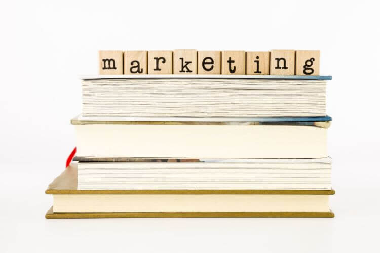 A stack of books with wooden blocks text "marketing" on top align  with white background.