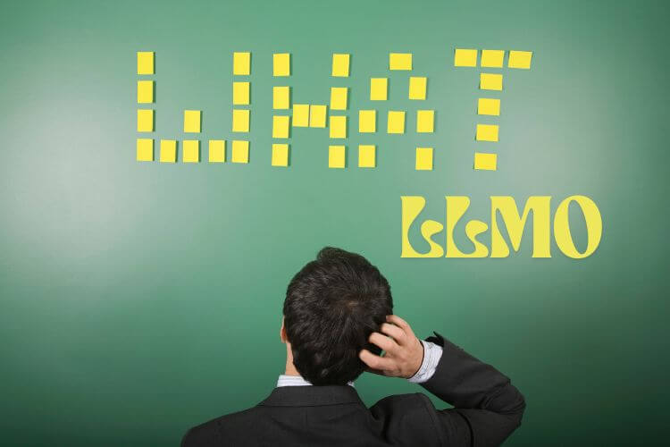 A man in a suit scratches his head while looking at a green board. Read the text "What LLMO"