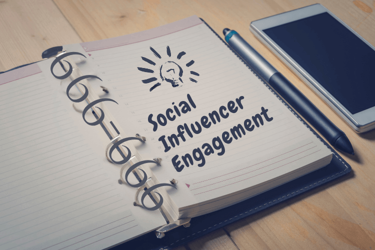 Notebook with handwritten text “Social Influencer Engagement” beside smartphone and pen on wooden desk.