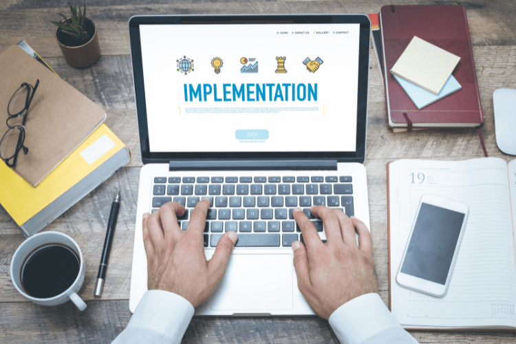 Person typing “Implementation” on a laptop screen surrounded by work essentials, symbolizing database rollout challenges